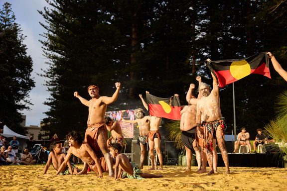 Dancers perform a traditional dance on sandy ground, holding Aboriginal flags, with trees and onlookers in the background.