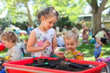 Two young girls gardening together at an outdoor event, using soil and tools in a red tray, surrounded by other children and adults.