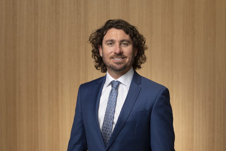 Councillor Adin Lang smiling at the camera with a brown background.