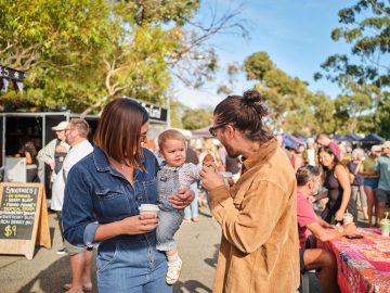 A man and woman lovingly hold a baby while exploring an outdoor market filled with vibrant stalls.