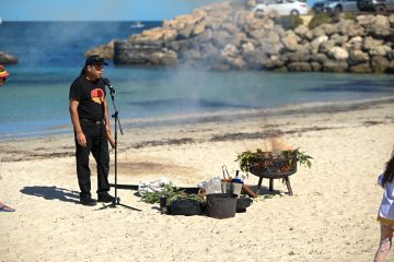 A man stands on a beach with a microphone, ready to perform against a scenic ocean backdrop.