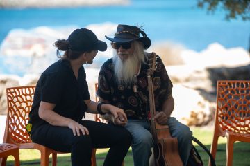 A man with a long beard sits on a chair beside a woman, both engaged in conversation.