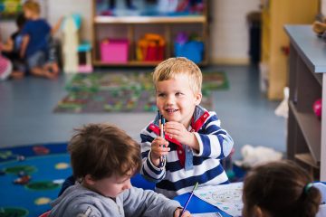 Happy children in a colourful classroom, a boy in striped shirt holds a marker, smiling. Another child colors beside him on a patterned blue rug.