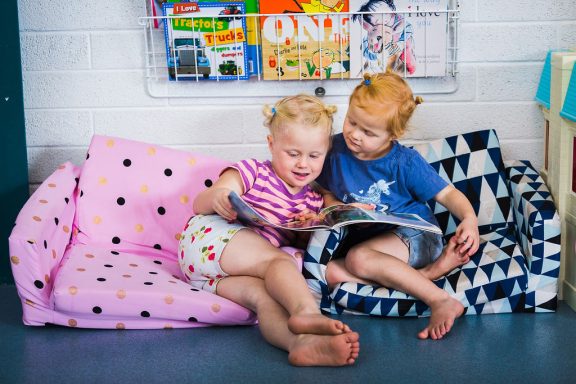 Two young children with similar hairstyles sitting together on colourful cushions, sharing a book. One wears purple stripes, the other a blue shirt. Children's books are displayed on a rack on the white brick wall behind them.
