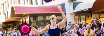 A street performer in a blue outfit juggles hats in front of a cheerful crowd outside shops, with people watching and taking photos on a sunny day.