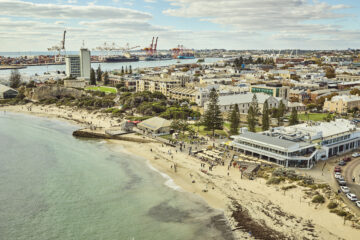 A coastal city scene with sandy beaches, people walking along the shore, buildings near the water, a busy port with cranes in the background, and scattered clouds in the sky.