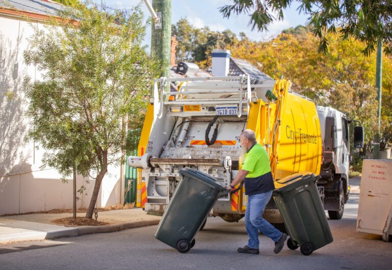 A sanitation worker in a neon green shirt moves two green trash bins toward a yellow garbage truck on a suburban street lined with trees and houses.