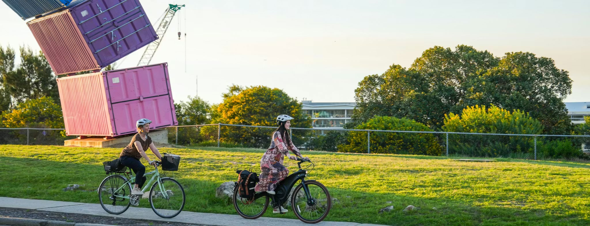 Two people ride bicycles on a path beside a grassy area with stacked pink shipping containers and a crane in the background, surrounded by trees and a building partially visible.