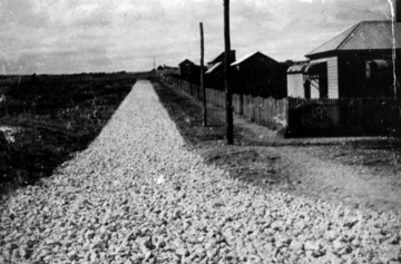 A black-and-white photo of a gravel road lined with utility poles, with wooden houses and a picket fence on the right side, and open land on the left under a cloudy sky.