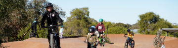 Several children wearing helmets ride bicycles on a dirt bike track surrounded by trees and greenery under a clear blue sky.