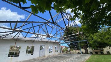 Partially demolished building with open walls, exposed steel roof beams, and no roof. Green trees and grass are visible outside, with a blue sky and clouds above.