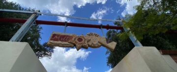 A decorative sign reading Mills Bakeries Park hangs between two metal posts against a blue sky with clouds and tree branches framing the view from below.