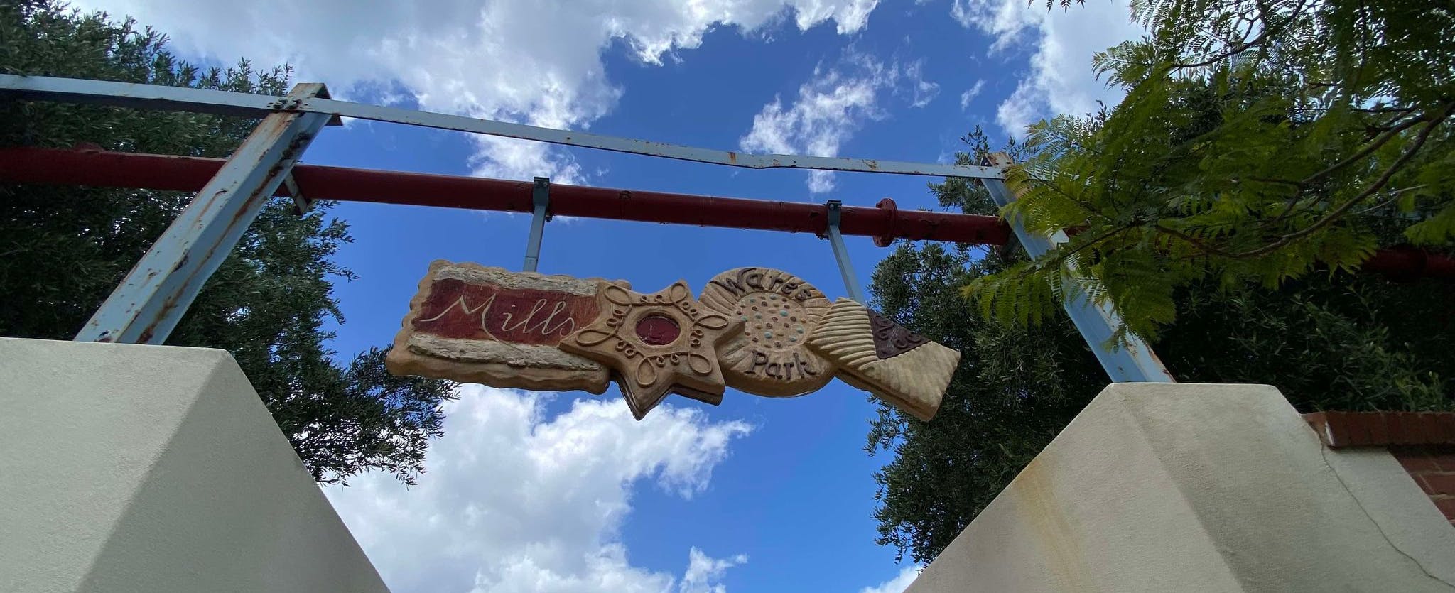 A decorative sign reading Mills Bakeries Park hangs between two metal posts against a blue sky with clouds and tree branches framing the view from below.