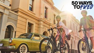 Two people ride bicycles on a sunny street lined with historic buildings and a vintage green car, with the text 50 Ways to Fred in the top right corner.