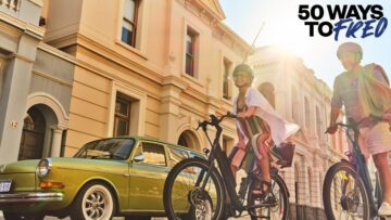 Two people wearing helmets ride bikes past a vintage green car and historic buildings, with sunlight streaming in. The text “50 Ways to FRED” appears in the top right corner.