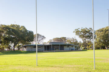 A sports field with two goalposts in the foreground, green grass, and a modern clubhouse building surrounded by trees under a clear blue sky.