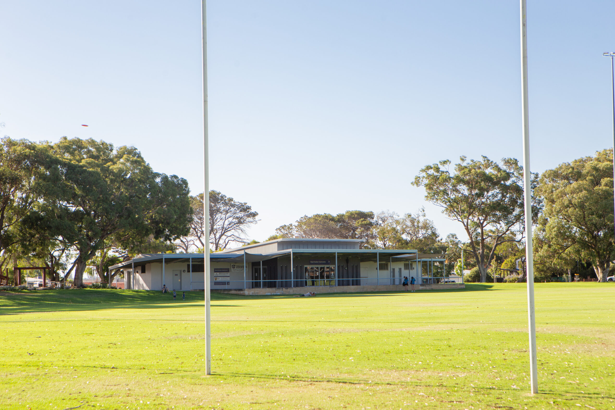 A sports field with two goalposts in the foreground, green grass, and a modern clubhouse building surrounded by trees under a clear blue sky.