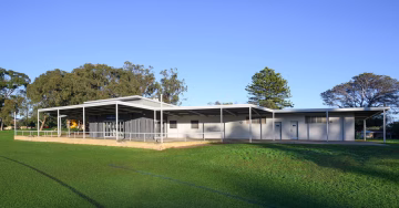 A modern, single-story sports pavilion with a large covered veranda overlooks a grassy field. Trees and a clear blue sky are in the background.