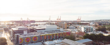 Aerial view of a city with colorful buildings in the foreground, shipping cranes, containers, and water in the background under a bright, cloudy sky.
